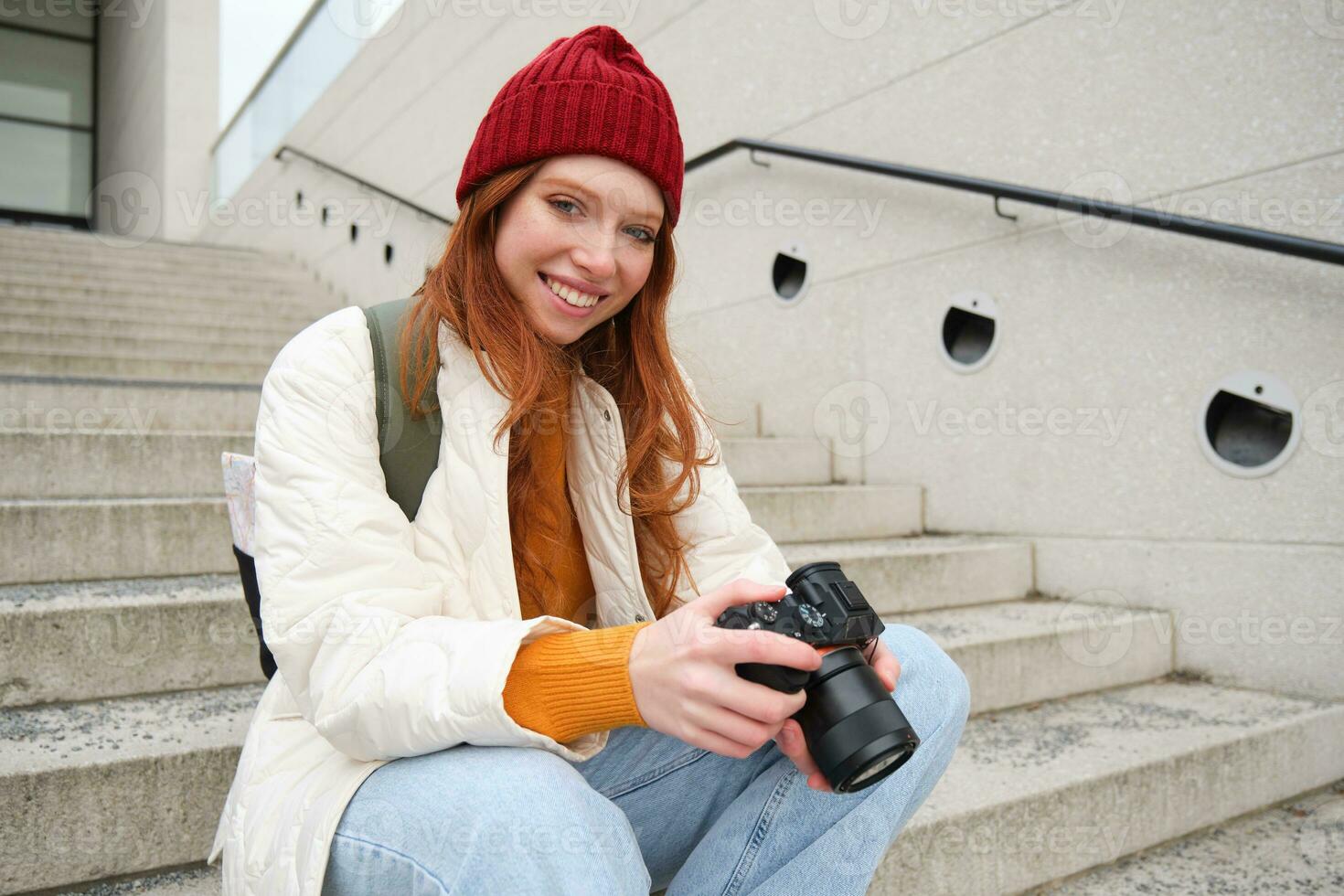 sonriente pelirrojo niña fotógrafo, cheques su tiros, sostiene cámara y mira a pantalla, toma fotos al aire libre, camina alrededor calle y lo hace estilo callejero disparo