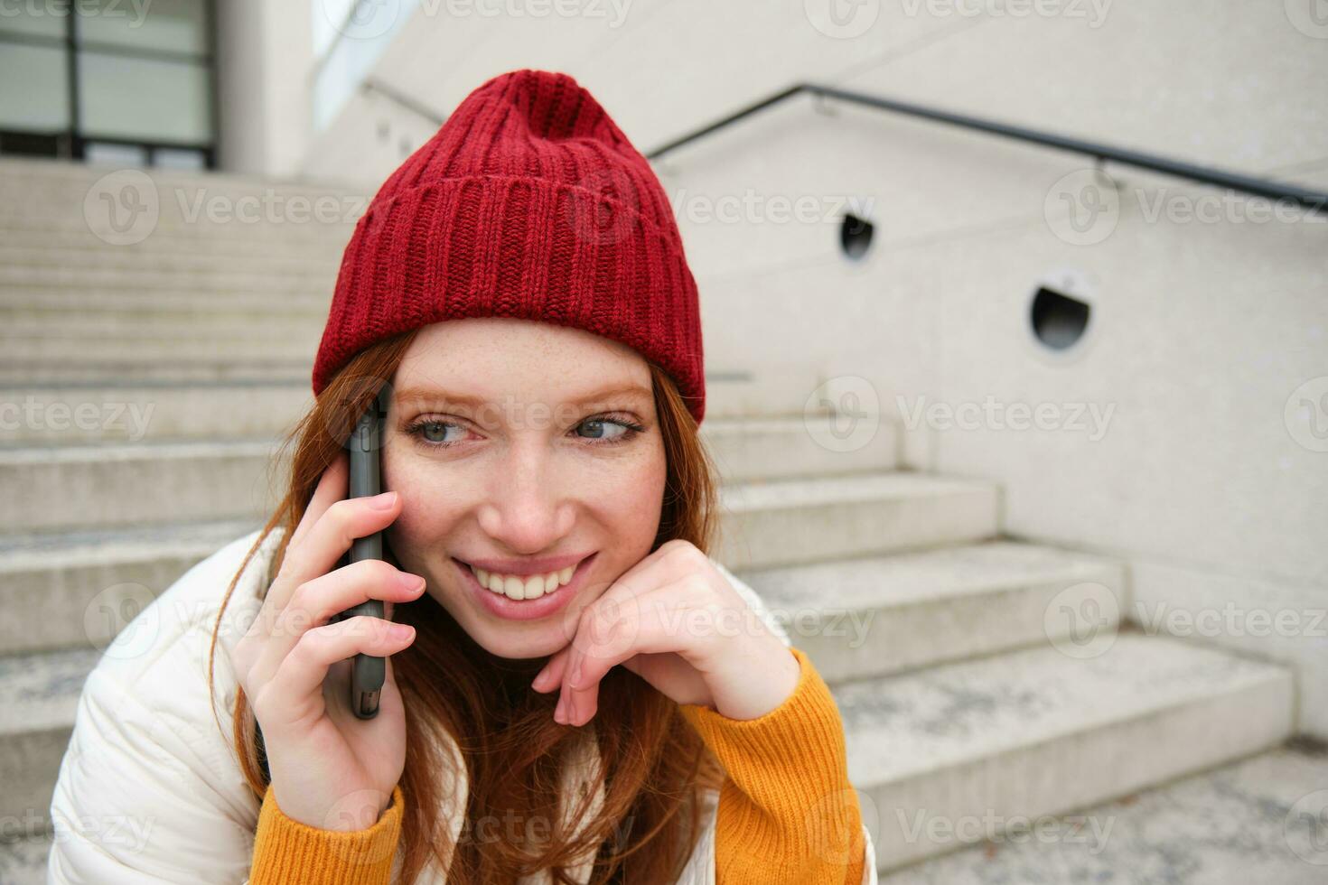 Beautiful smiling redhead female model, sits on street and talks on mobile phone, uses smartphone app to call abroad, laughing during telephone conversation photo