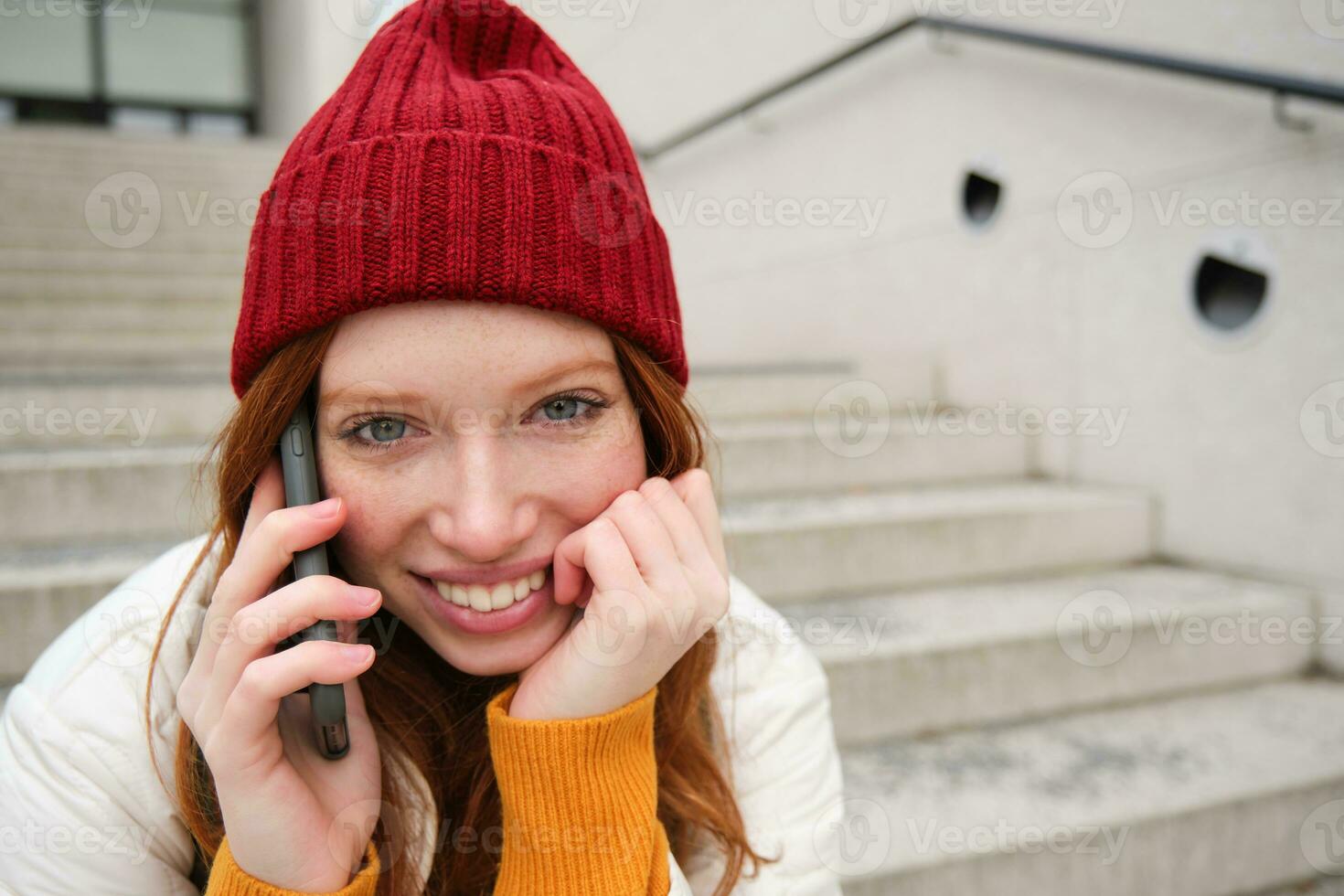 joven elegante pelirrojo niña en rojo sombrero, se sienta en calle y negociaciones en móvil teléfono, tiene teléfono conversación, anillos su amigo mientras relaja al aire libre foto