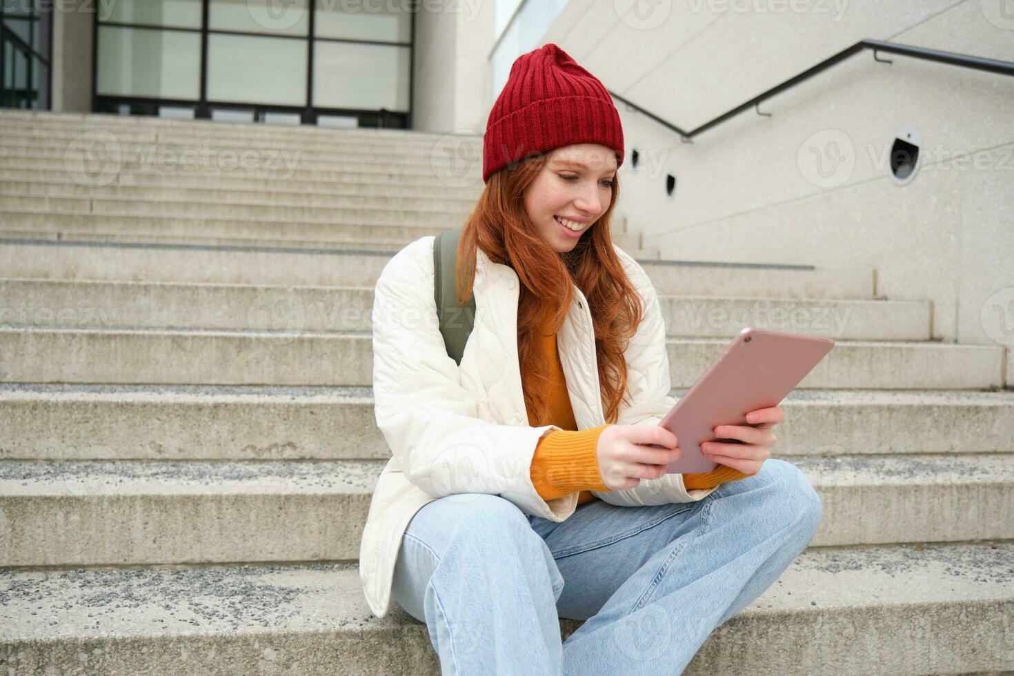 Young stylish girl, redhead female students sits on stairs outdoors with digital tablet, reads, uses social media app on gadget, plays games while waits on street photo