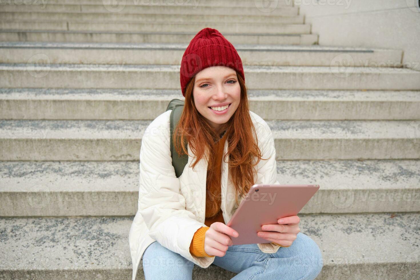 Happy stylish redhead girl, student in red hat, holds digital tablet, uses social media app, searches something online, connects to wifi photo