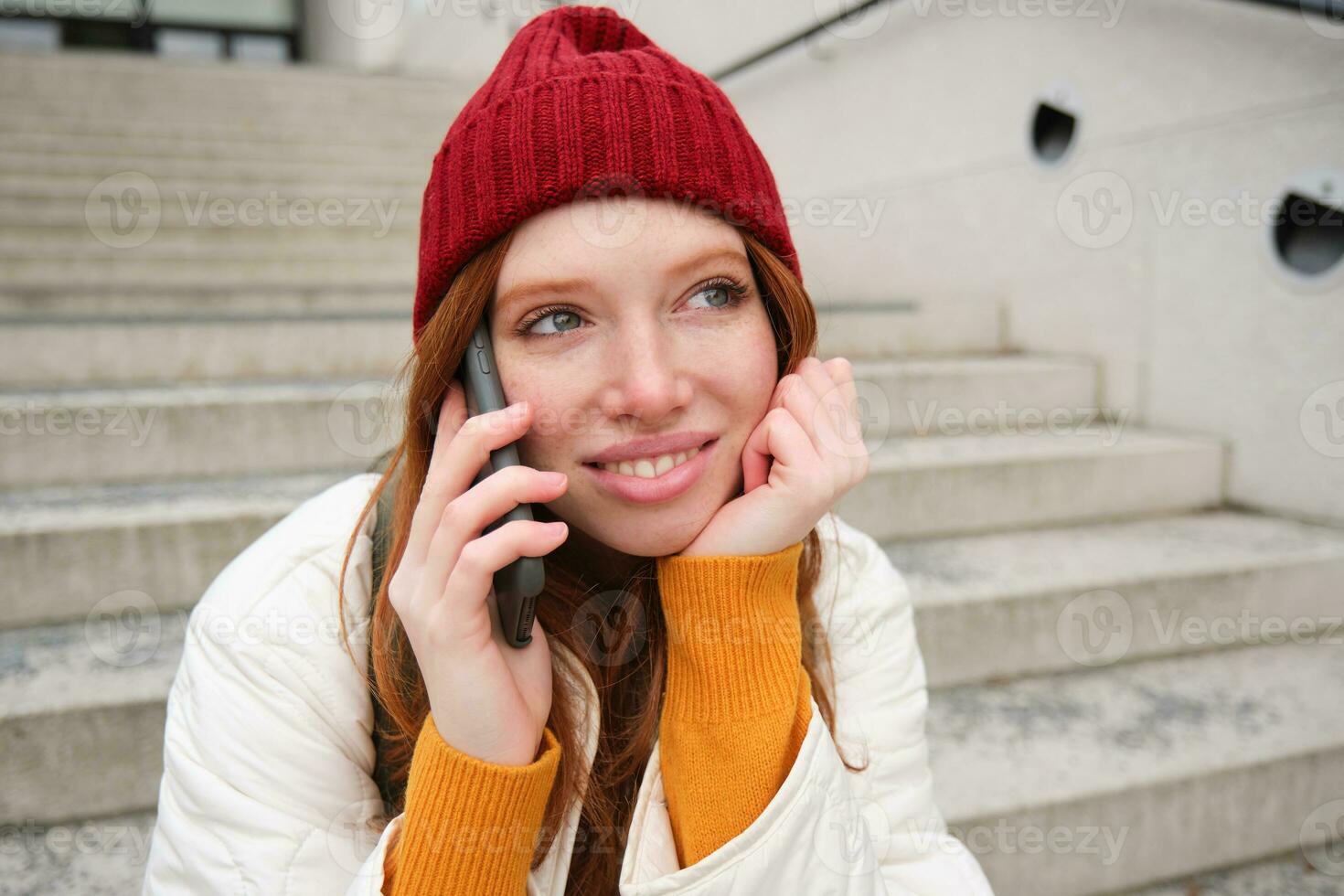 hermosa sonriente pelirrojo hembra modelo, se sienta en calle y negociaciones en móvil teléfono, usos teléfono inteligente aplicación a llamada en el extranjero, riendo durante teléfono conversacion foto