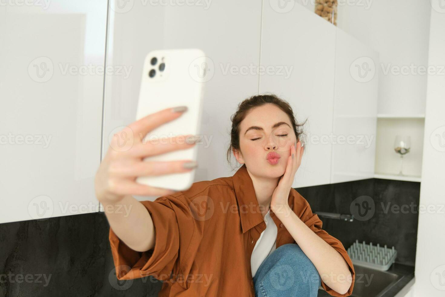 Young charismatic girl takes selfies in the kitchen, posing for photo, holds mobile phone with extended hand photo