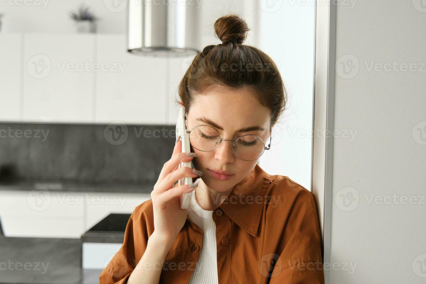 Portrait of young sad woman in glasses, talking on mobile phone, listening to voice message on smartphone with upset, concerned face expression, standing at home, leaning on wall photo