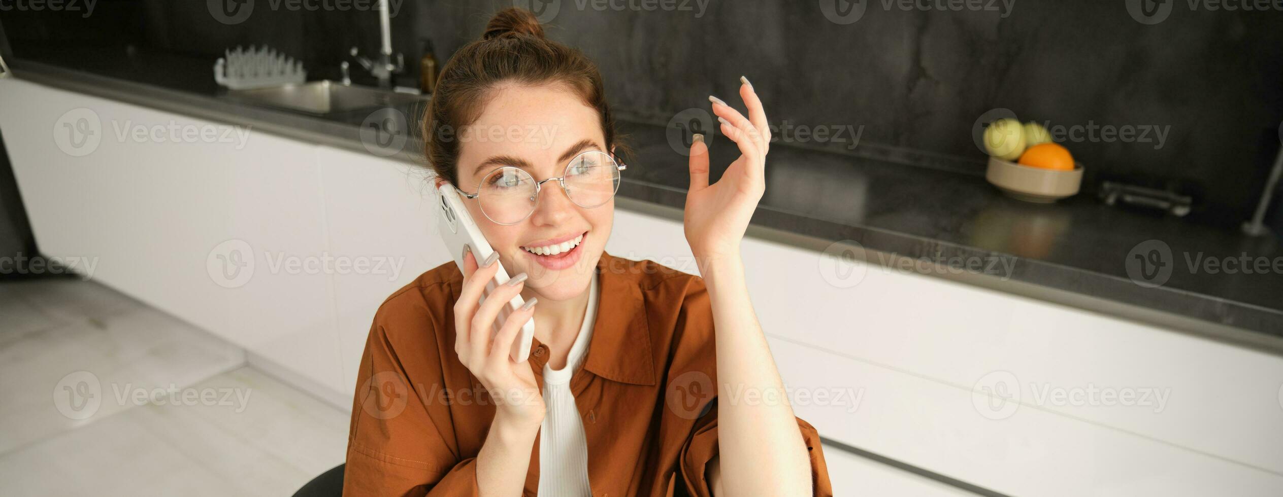 emocionado joven mujer hablando en móvil teléfono en frente de computadora portátil, sentado en cocina con contento cara expresión, teniendo un conversacion foto