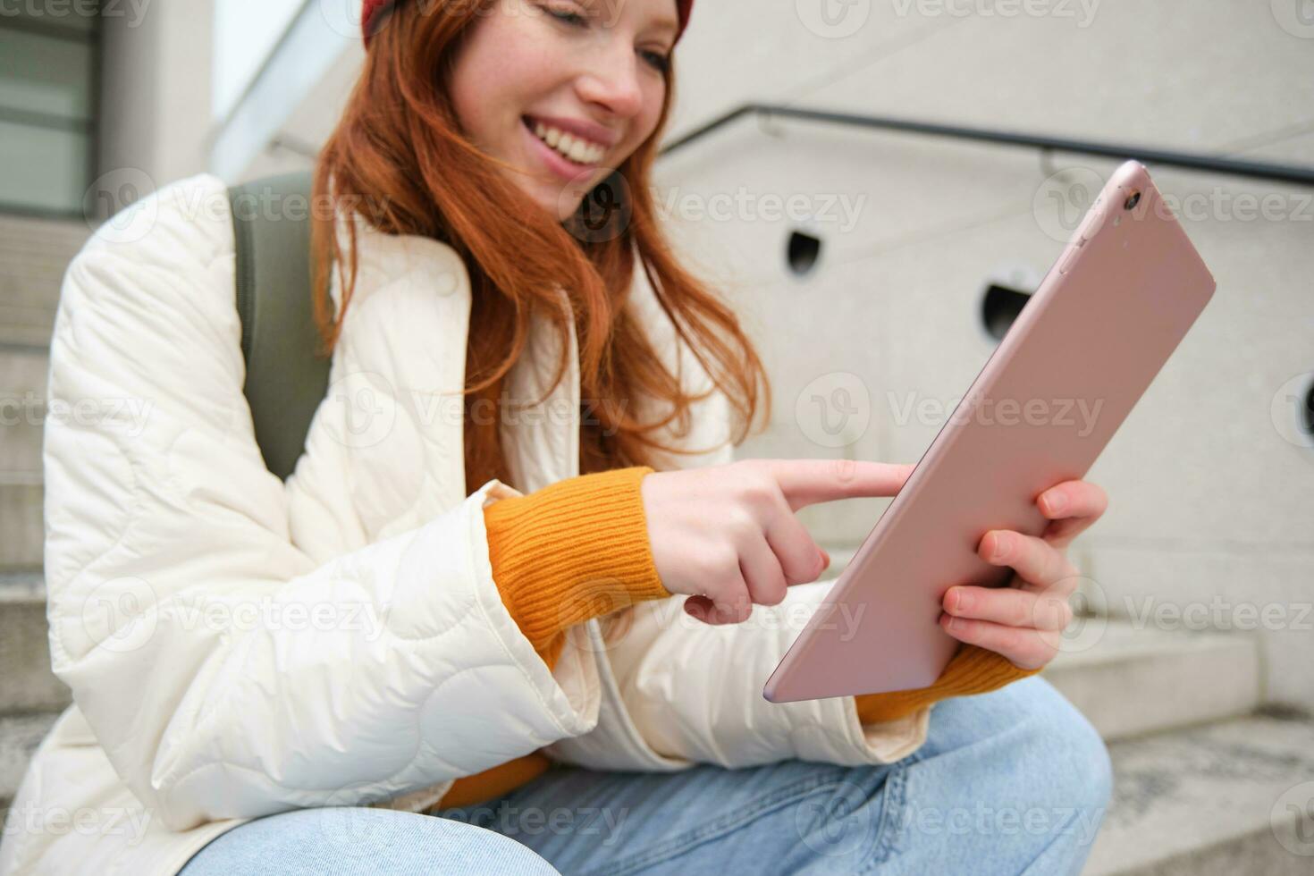 Happy stylish redhead girl, student in red hat, holds digital tablet, uses social media app, searches something online, connects to wifi photo