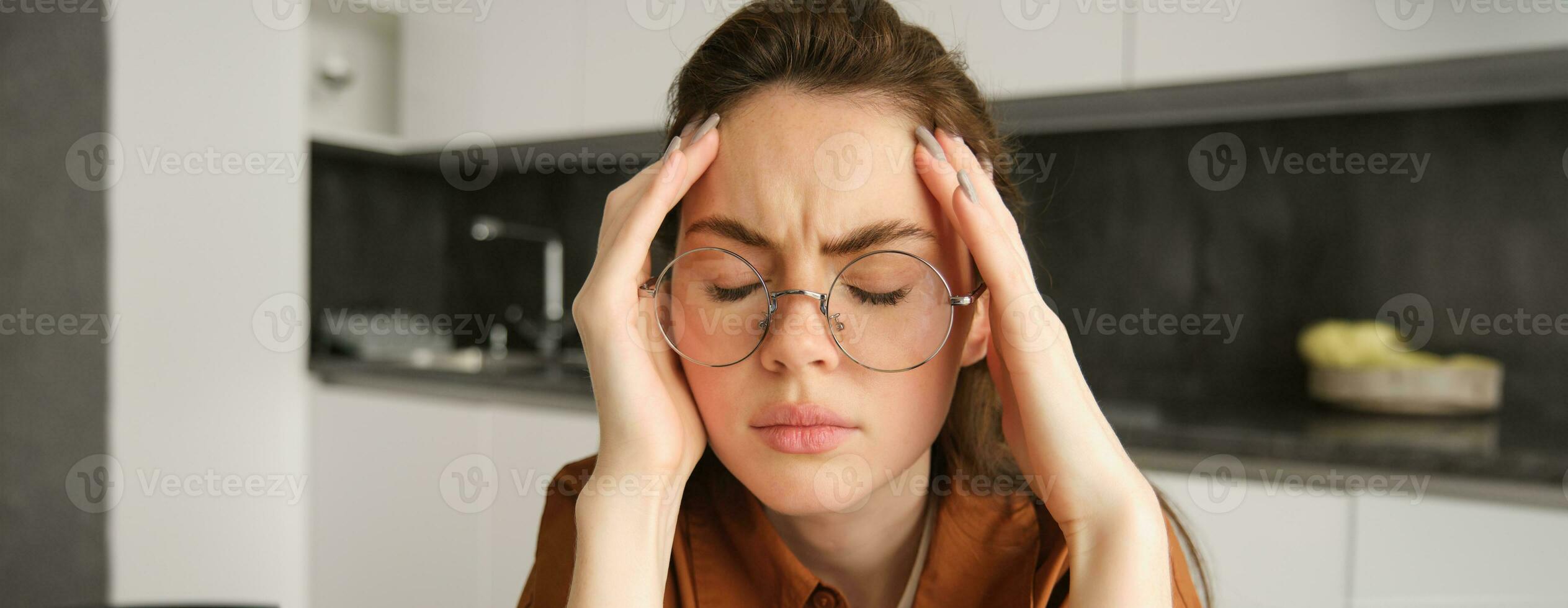 retrato de cansado joven trabajando mujer en anteojos, sostiene manos en cabeza, sufre doloroso migraña, tiene dolor en templos, sentado con dolor de cabeza en cocina foto