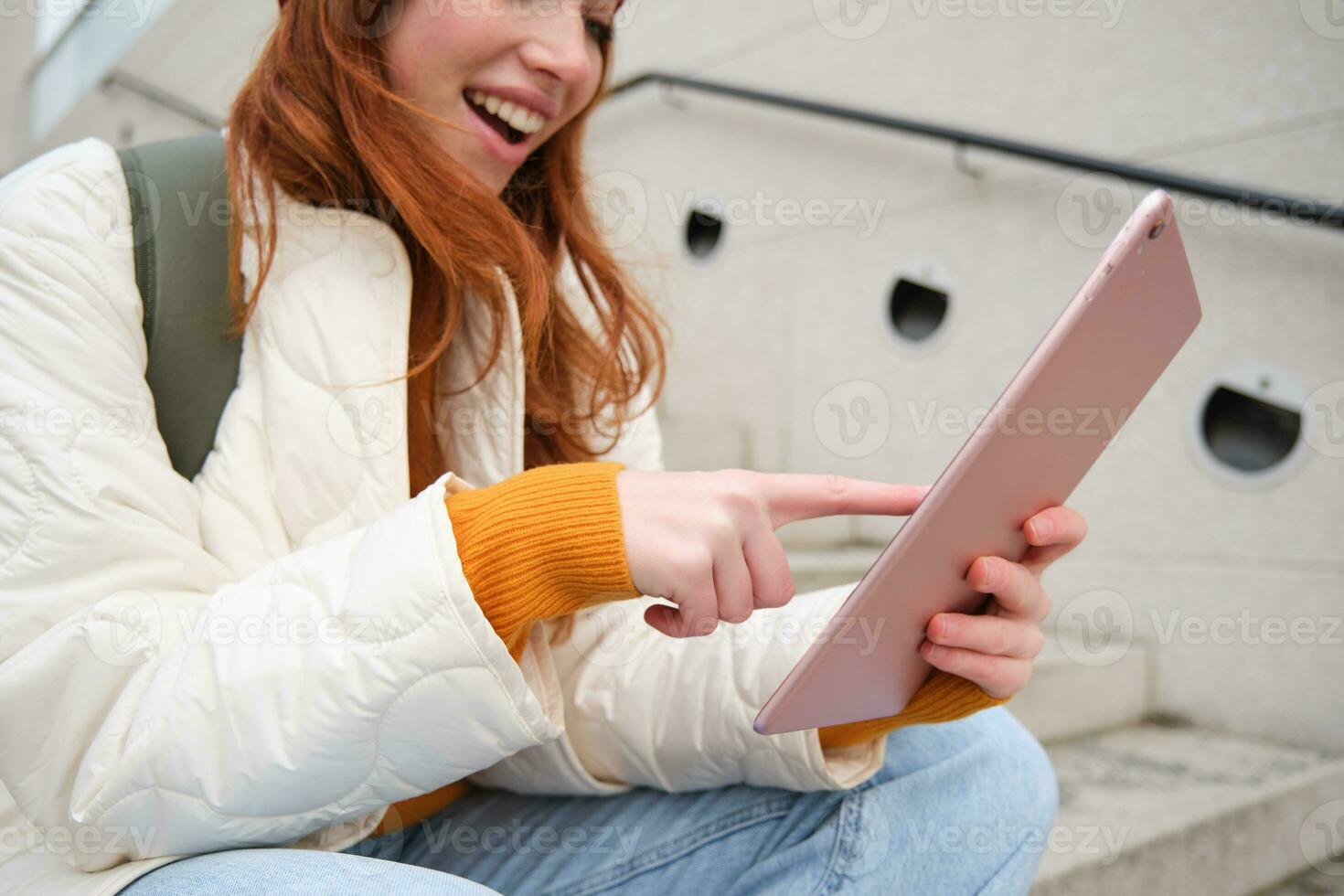 Young stylish girl, redhead female students sits on stairs outdoors with digital tablet, reads, uses social media app on gadget, plays games while waits on street photo