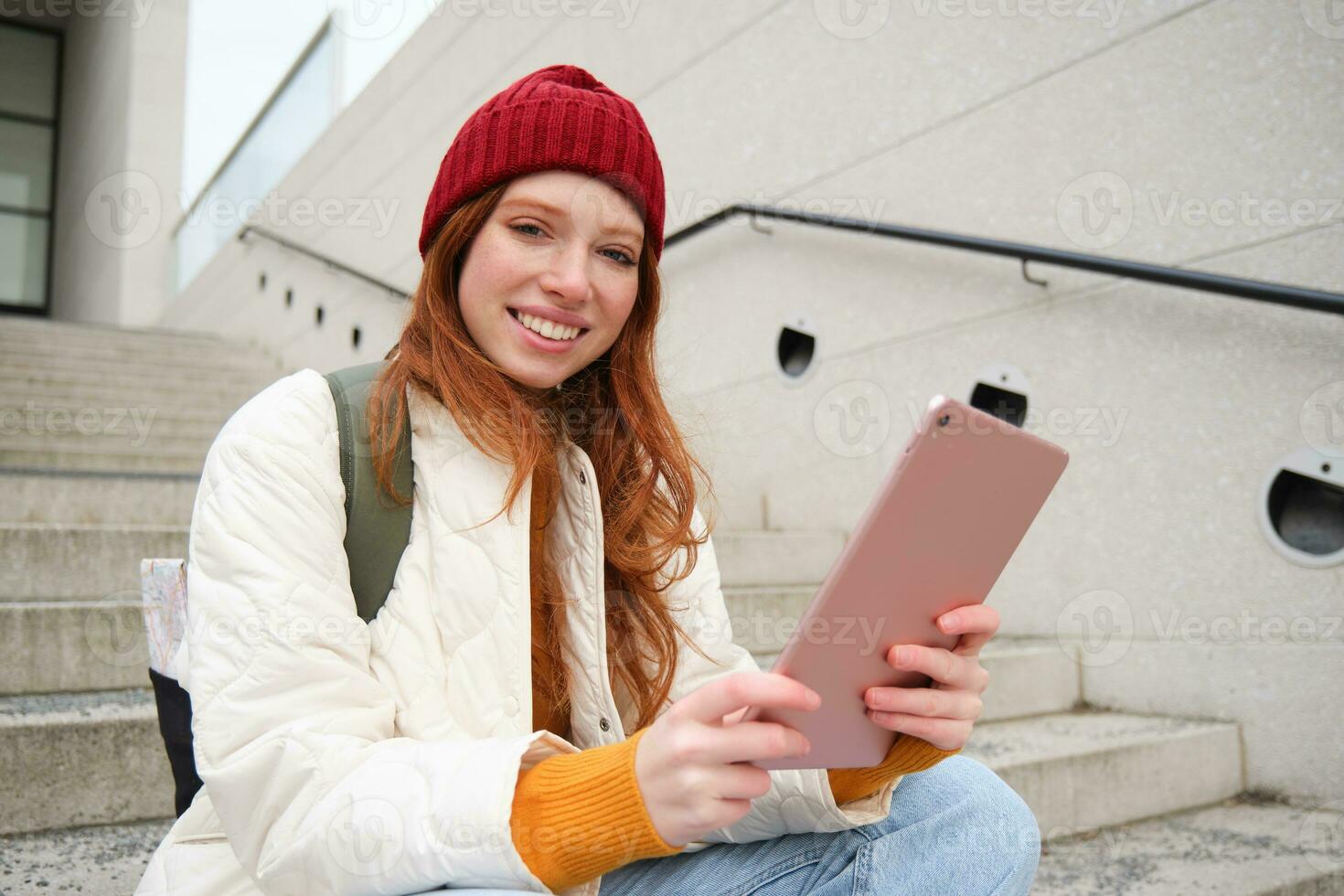 Happy stylish redhead girl, student in red hat, holds digital tablet, uses social media app, searches something online, connects to wifi photo