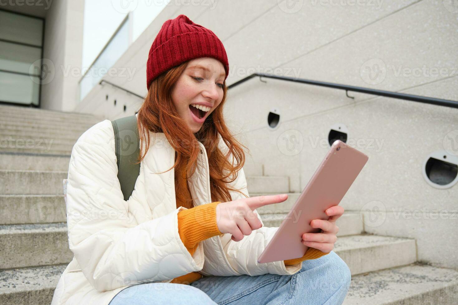 Young stylish girl, redhead female students sits on stairs outdoors with digital tablet, reads, uses social media app on gadget, plays games while waits on street photo