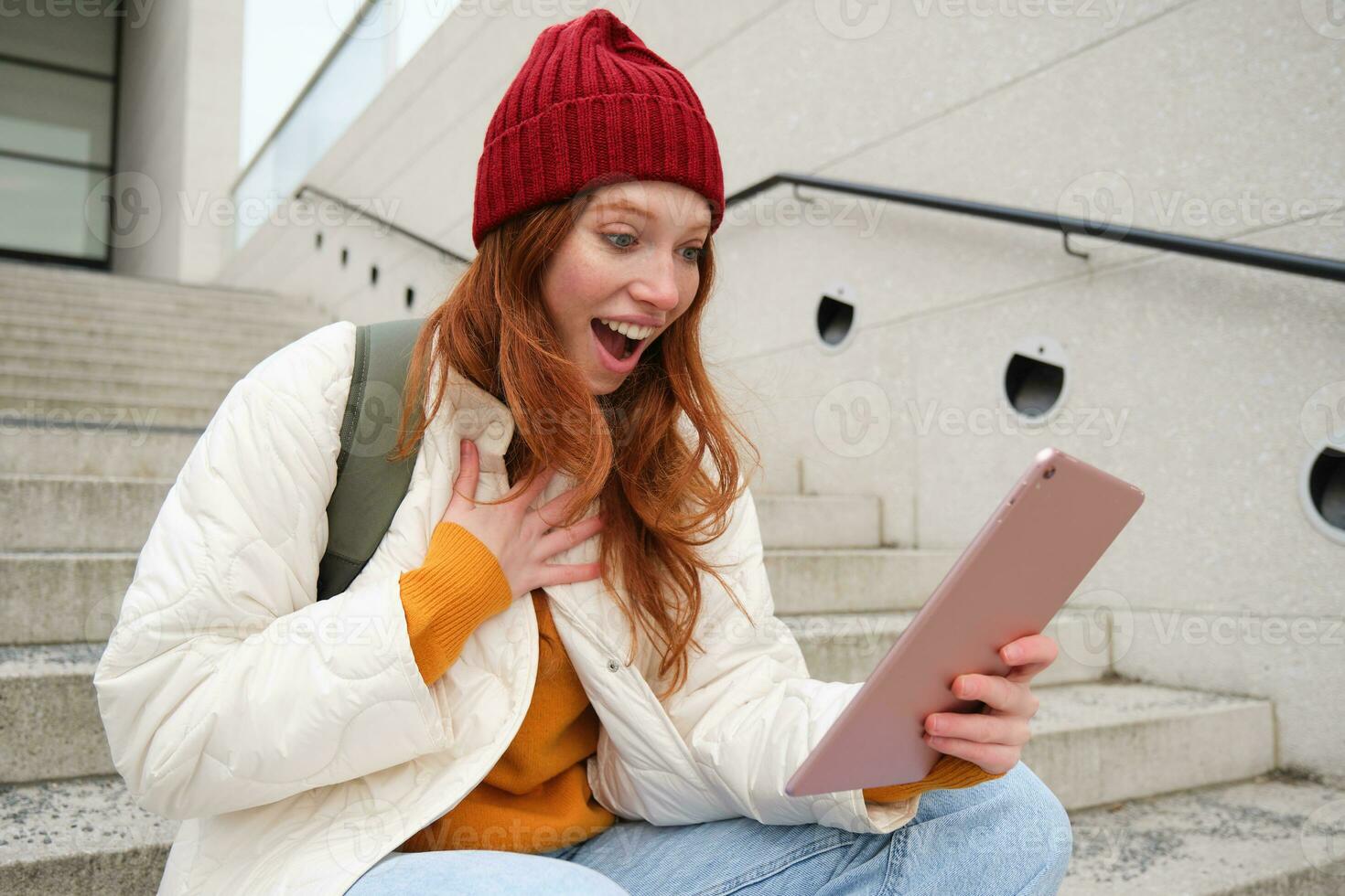 Happy stylish redhead girl, student in red hat, holds digital tablet, uses social media app, searches something online, connects to wifi photo