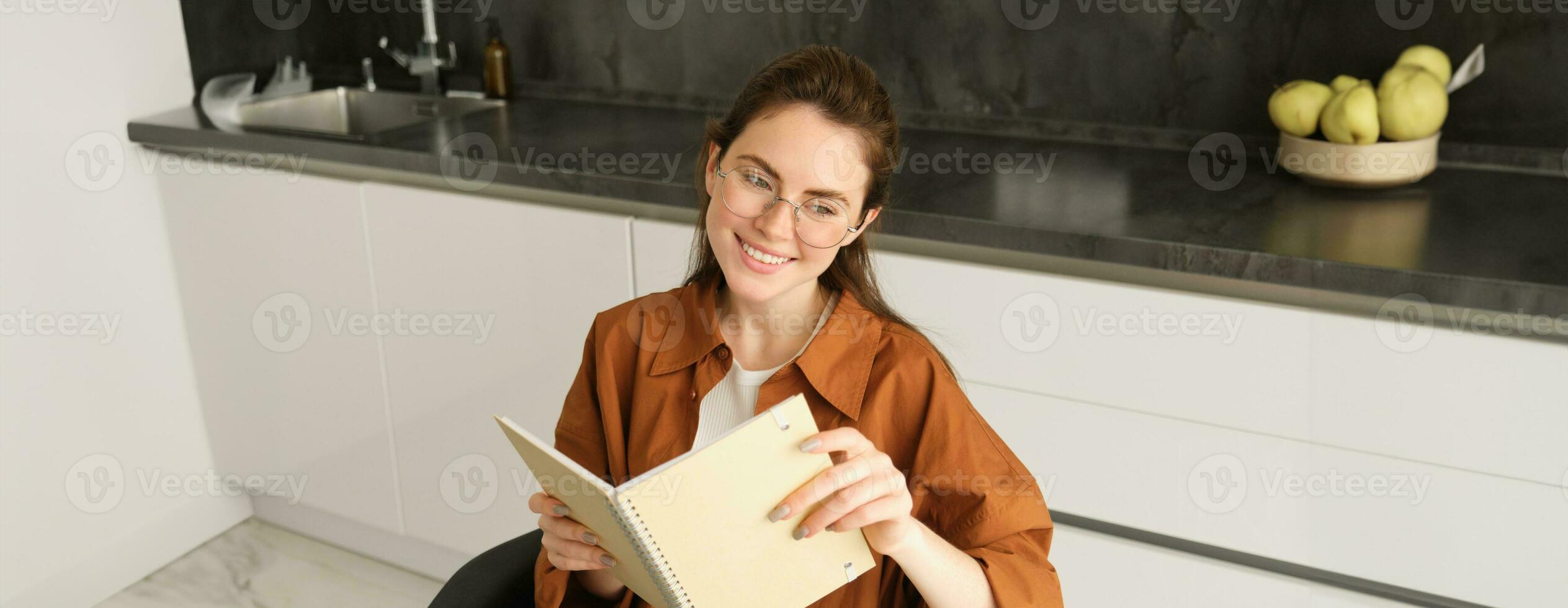 Close up portrait of young woman, student in kitchen, holding notebook, revising for exam at home, studying, reading her planner photo