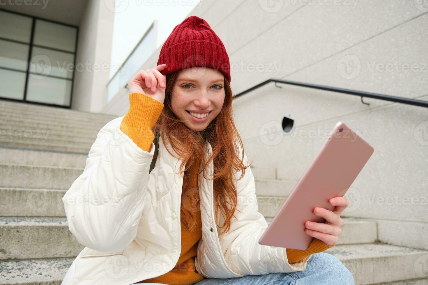 Young stylish girl, redhead female students sits on stairs outdoors with digital tablet, reads, uses social media app on gadget, plays games while waits on street photo
