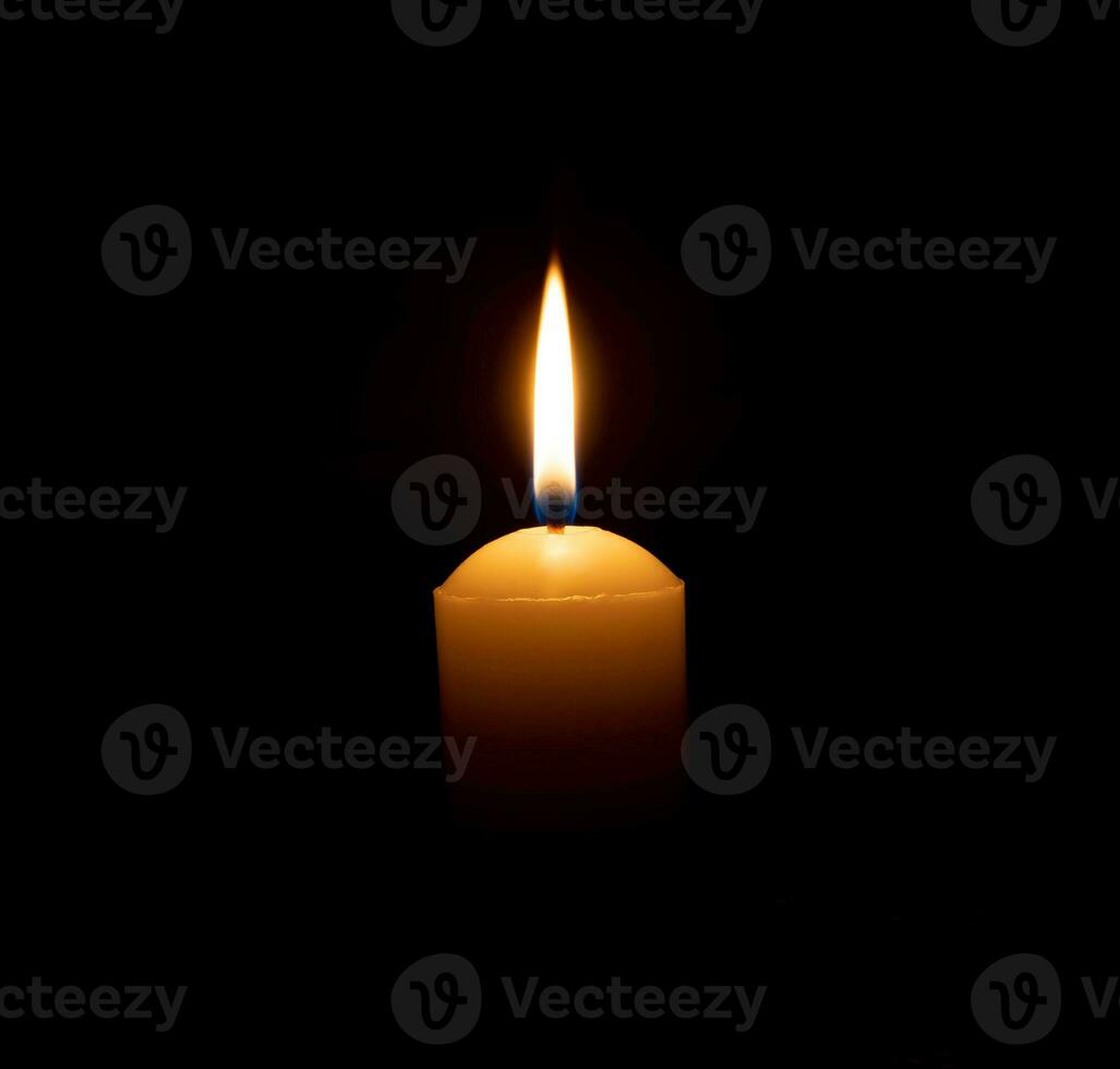 A single burning candle flame or light glowing on a big yellow candle on black or dark background on table in church for Christmas, funeral or memorial service with copy space photo