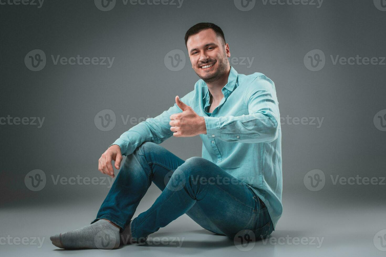 Appealing casual young man sitting on the floor, looking to the camera smiling photo