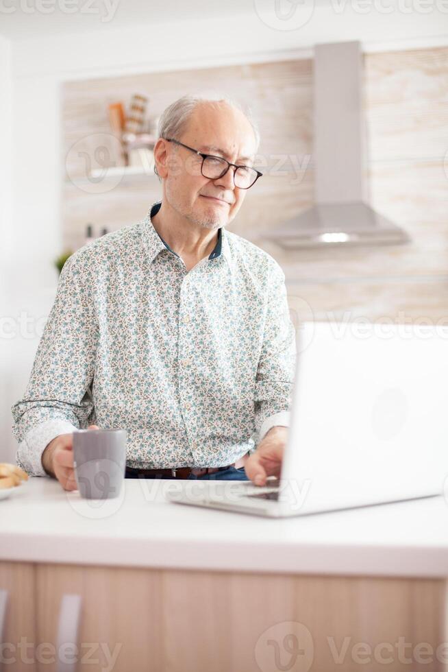 Grayhaired senior retired man browsing on internet. Daily life of senior man in kitchen during breakfast using laptop holding a cup of coffee. Elderly retired person working from home, telecommuting using remote internet job online communication photo