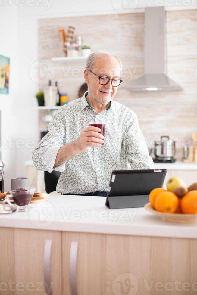 Elderly man surfing on internet using tablet computer during breakfast in kitchen. Senior person with tablet portable pad PC in retirement age using mobile apps, modern internet online information technology with touchscreen photo
