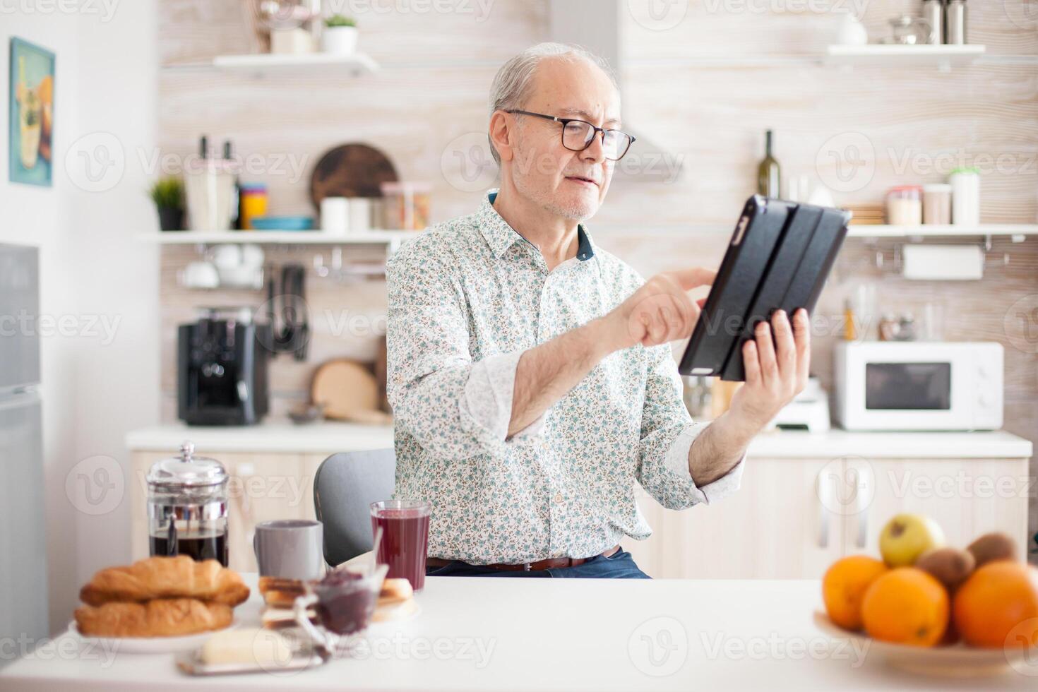 Happy elderly person with portable tablet PC in kitchen during breakfast. Senior person with tablet portable pad PC in retirement age using mobile apps, modern internet online information technology with touchscreen photo