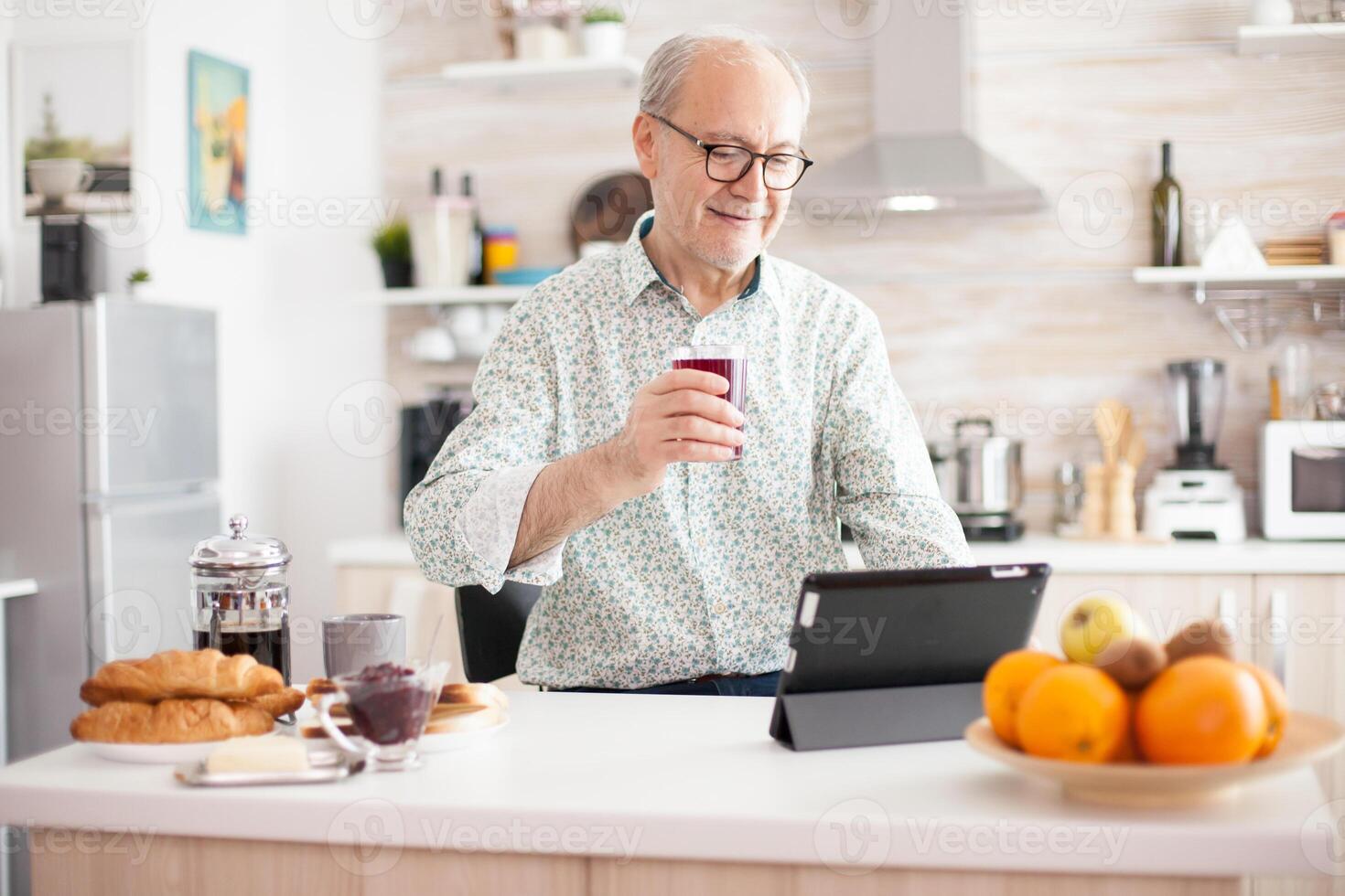 Elderly man using tablet pc in kitchen during breakfast wearing glasses. Elderly person with tablet portable pad PC in retirement age using mobile apps, modern internet online information technology with touchscreen photo