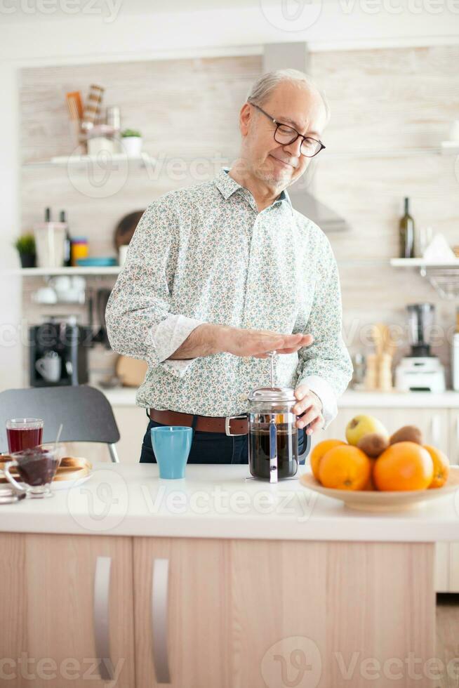 Senior man making coffee using french press during breakfast in kitchen. Elderly person in the morning enjoying fresh brown cafe espresso cup caffeine from vintage mug, filter relax refreshment photo