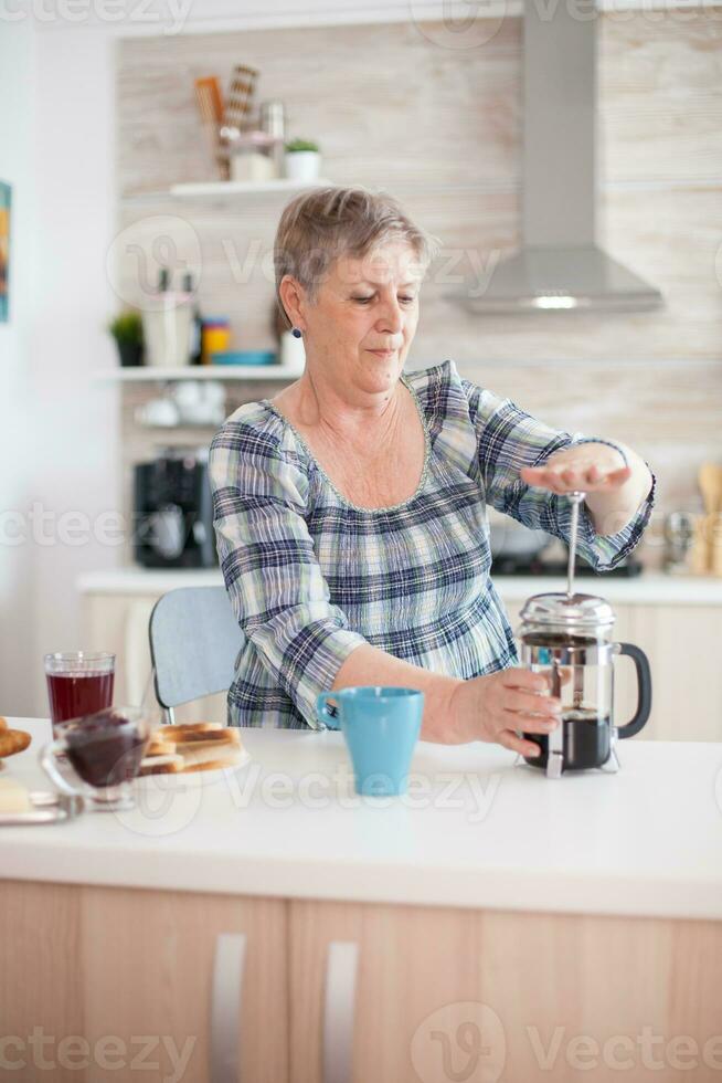 Senior woman pressing on french press to make tasty coffee during breakfast in kitchen. Elderly person in the morning enjoying fresh brown cafe espresso cup caffeine from vintage mug, filter relax refreshment photo