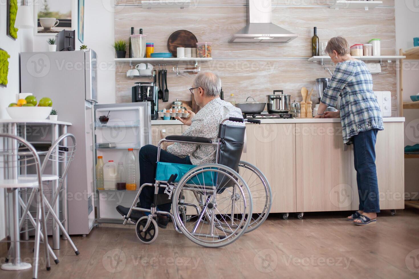 Disabled senior man in wheelchair taking eggs carton from refrigerator for wife in kitchen. Senior woman helping handicapped husband. Living with disabled person with walking disabilities photo
