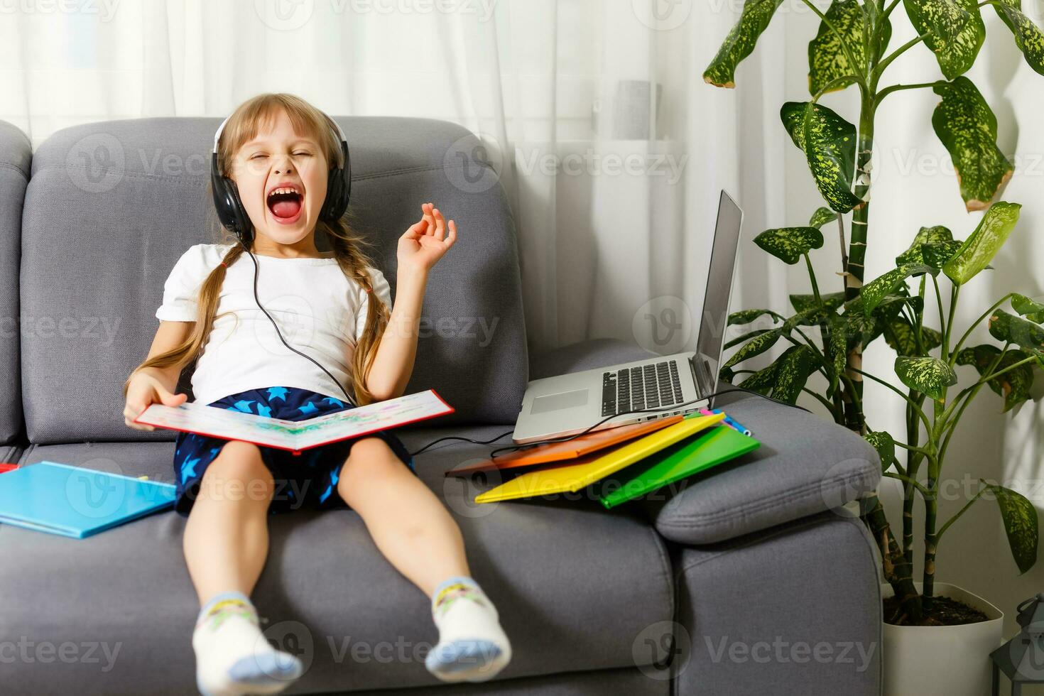 Photo of little pretty pupil school lady turn on skype laptop notebook online lesson hold open book ready answer teacher questions sit sofa distance quarantine study comfort room indoors