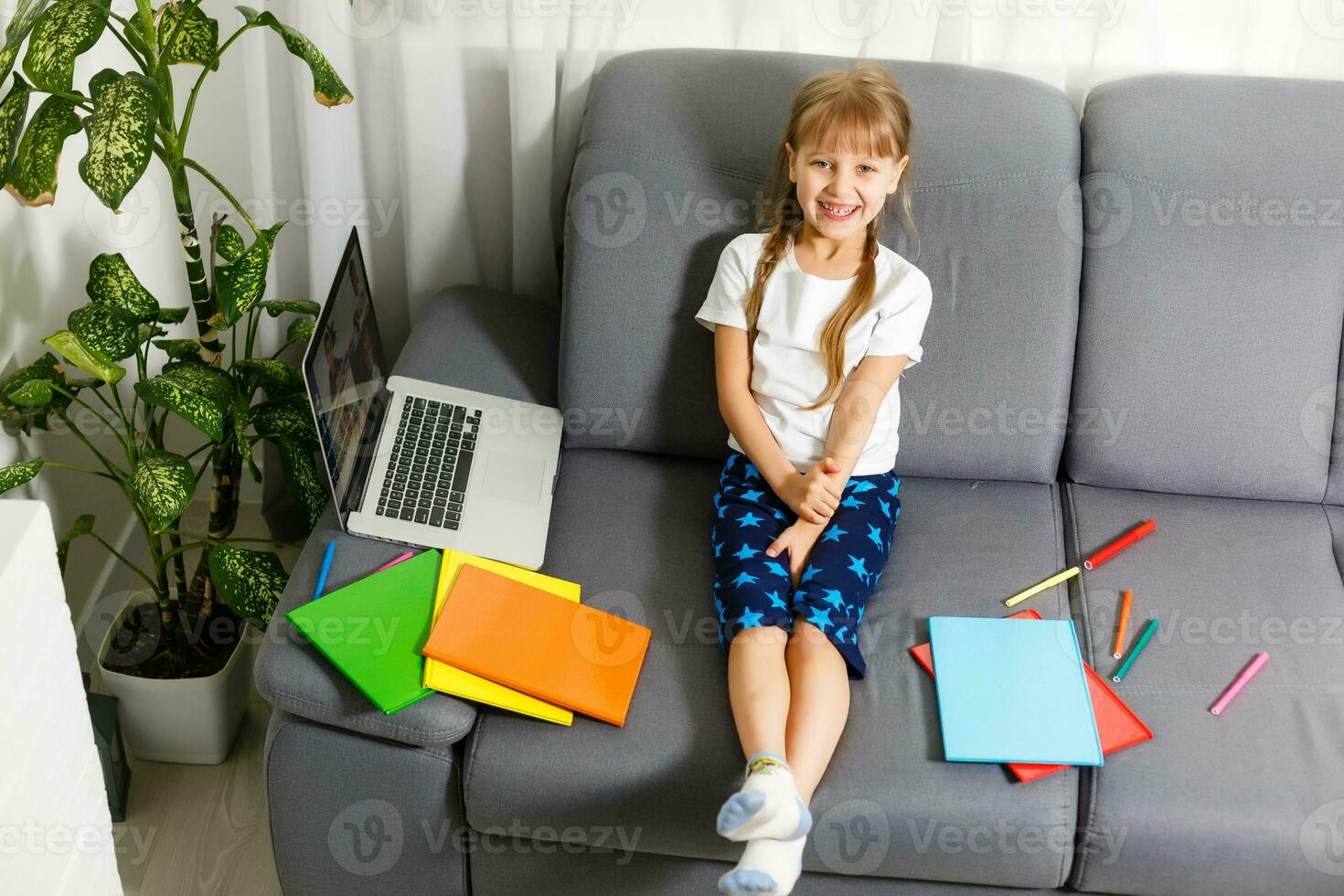 Photo of little pretty pupil school lady turn on skype laptop notebook online lesson hold open book ready answer teacher questions sit sofa distance quarantine study comfort room indoors