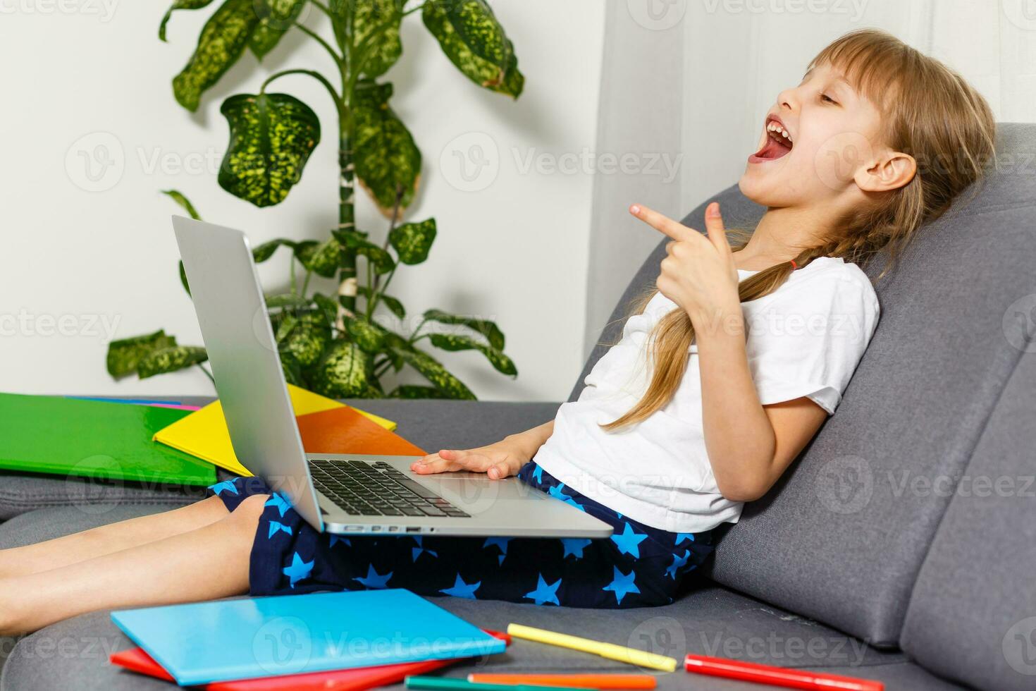 Photo of little pretty pupil school lady turn on skype laptop notebook online lesson hold open book ready answer teacher questions sit sofa distance quarantine study comfort room indoors