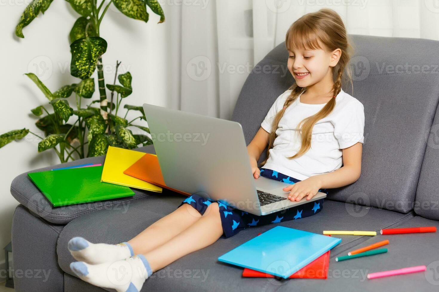 Photo of little pretty pupil school lady turn on skype laptop notebook online lesson hold open book ready answer teacher questions sit sofa distance quarantine study comfort room indoors