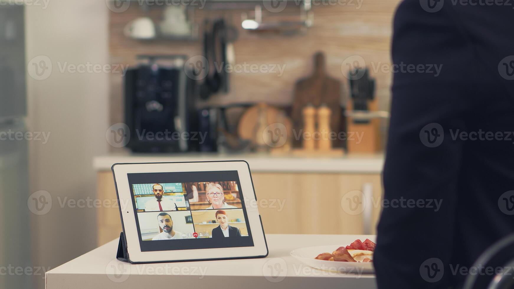 Business woman using smart device while eating breakfast in kitchen. Young freelancer at home talking on a video call with her colleagues from the office, using modern internet technology photo
