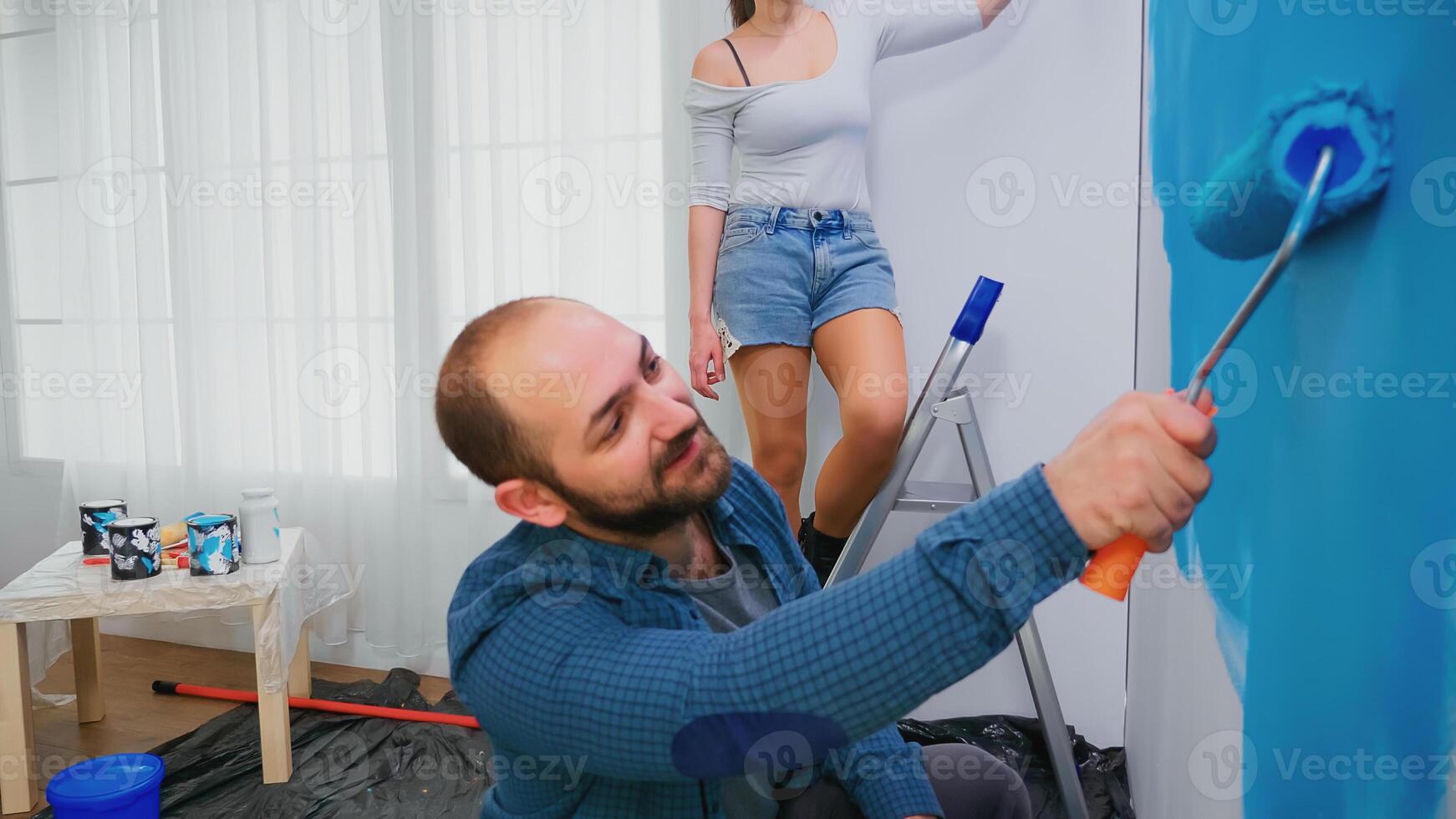 Wife painting wall on ladder during home renovation using roller brush. Couple redecorating. Home decoration and renovation in cozy apartment flat, repair and makeover photo