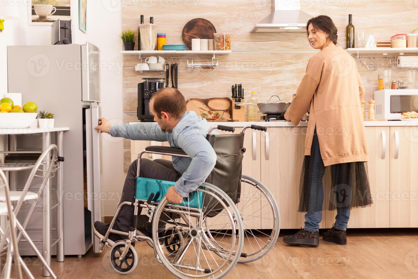Woman in kitchen looking at husband with walking disability trying to open refrigerator door. Disabled paralyzed handicapped man with walking disability integrating after an accident. photo