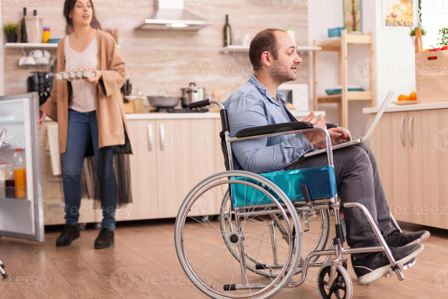 Disabled man in wheelchair during video call on laptop in kitchen while is holding box of eggs and holding refrigerator door open. Disabled paralyzed handicapped man with walking disability integrating after an accident. photo