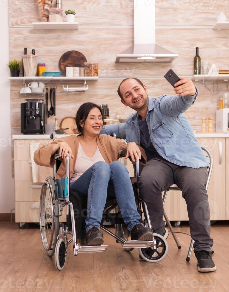 Positive woman in wheelchair and husband taking a selfie in kitchen using smartphone. Disabled paralyzed handicapped woman with walking disability integrating after an accident. photo
