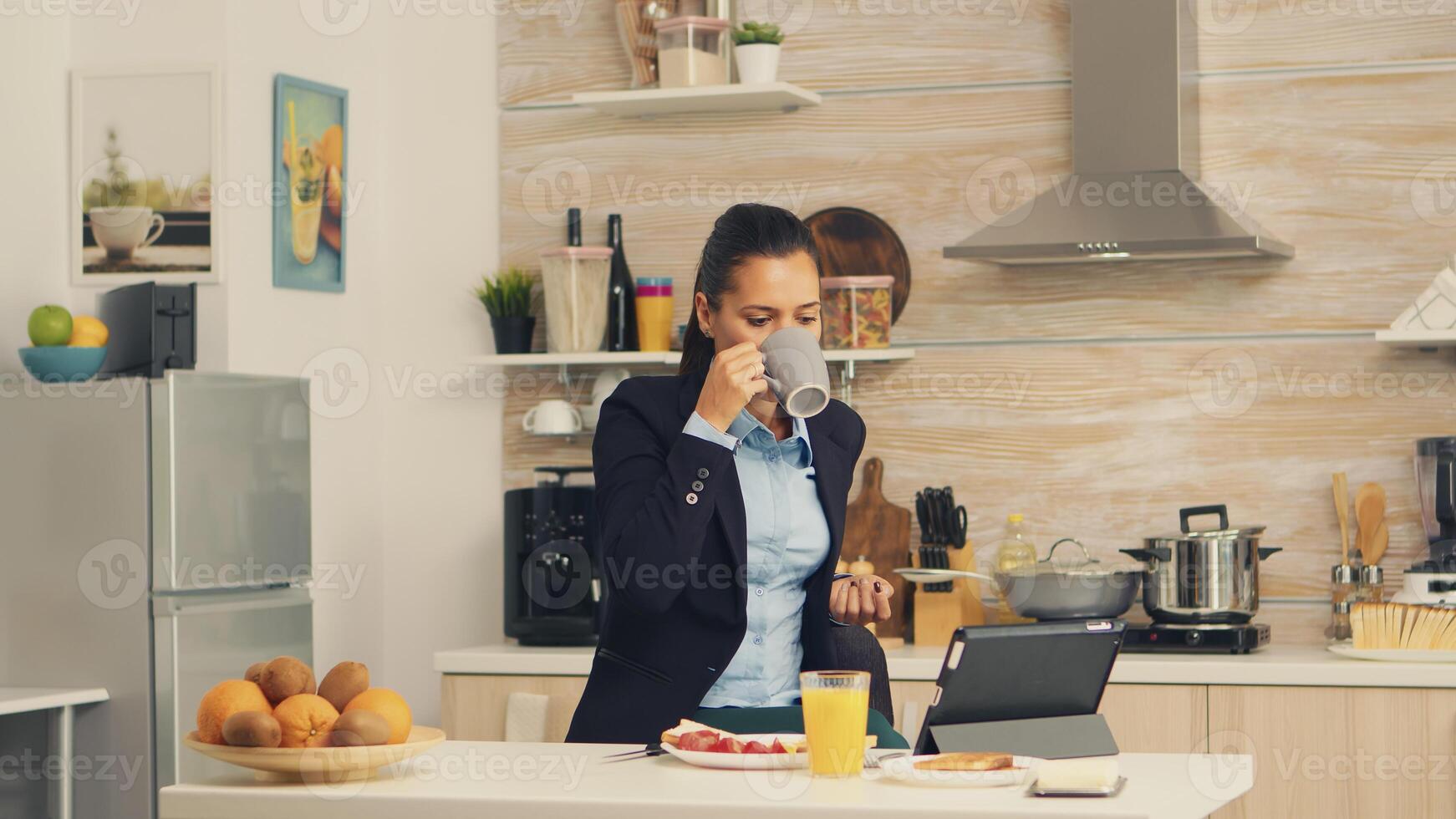 Freelancer drinking coffee in the morning on table top during breakfast using tablet computer. Business woman reading the last news online before going to work, using modern technology in the kitchen while eating a healthy meal photo