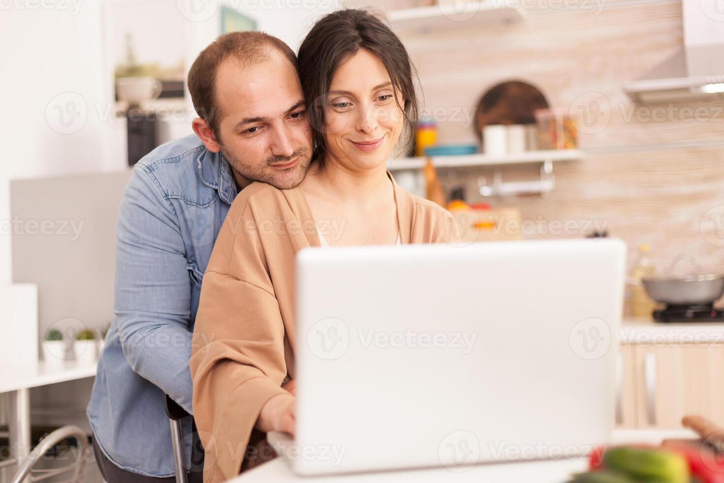 Smiling couple using laptop in kitchen. Man hugging wife. Happy loving cheerful romantic in love couple at home using modern wifi wireless internet technology photo