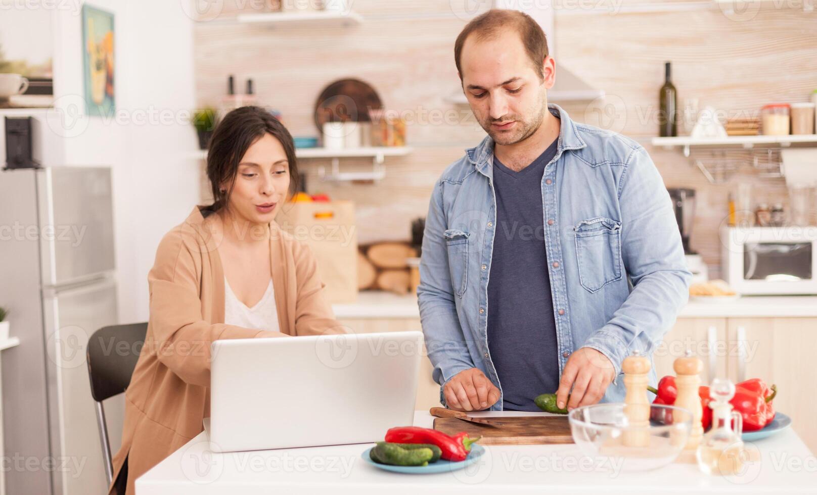 Couple prepares healthy salad looking at online recipe on laptop in kitchen. Man helping woman to prepare healthy organic dinner, cooking together. Romantic cheerful love relationship photo