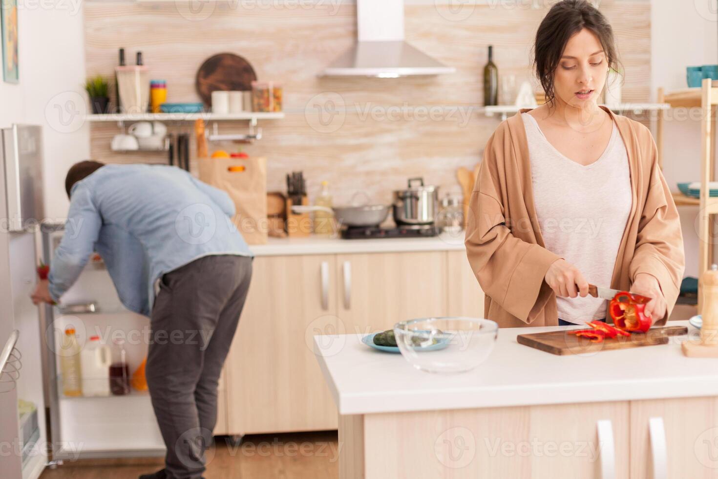 Husband looking inside of refrigerator and wife is cutting red bell pepper in kitchen using sharp knife. Funny happy in love couple at home spending time together, cooking healthy and smiling photo