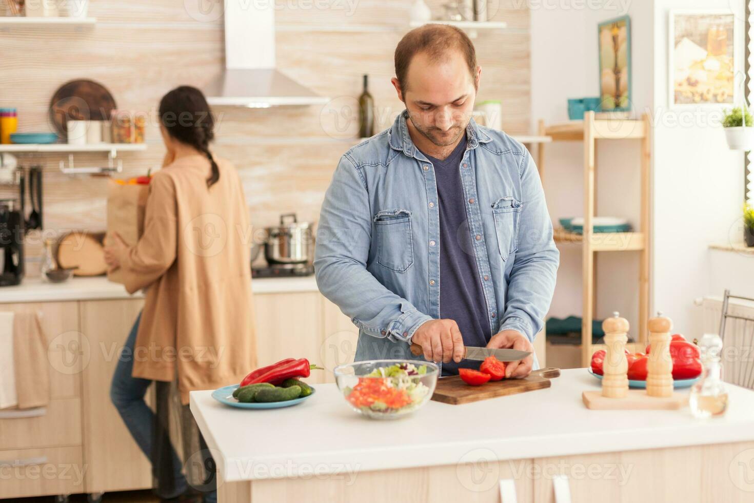 Husband chopping tomatoes for salad on wooden cutting board in kitchen. Wife holding paper groceries bag. Happy in love cheerful and carefree couple helping each other to prepare meal photo