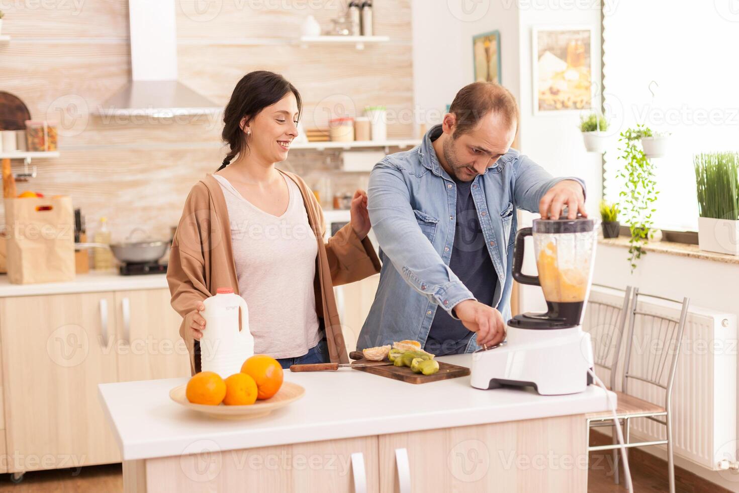 Cheerful couple prepares smoothie using blender. Wife holding milk bottle in kitchen. Healthy carefree and cheerful lifestyle, eating diet and preparing breakfast in cozy sunny morning photo