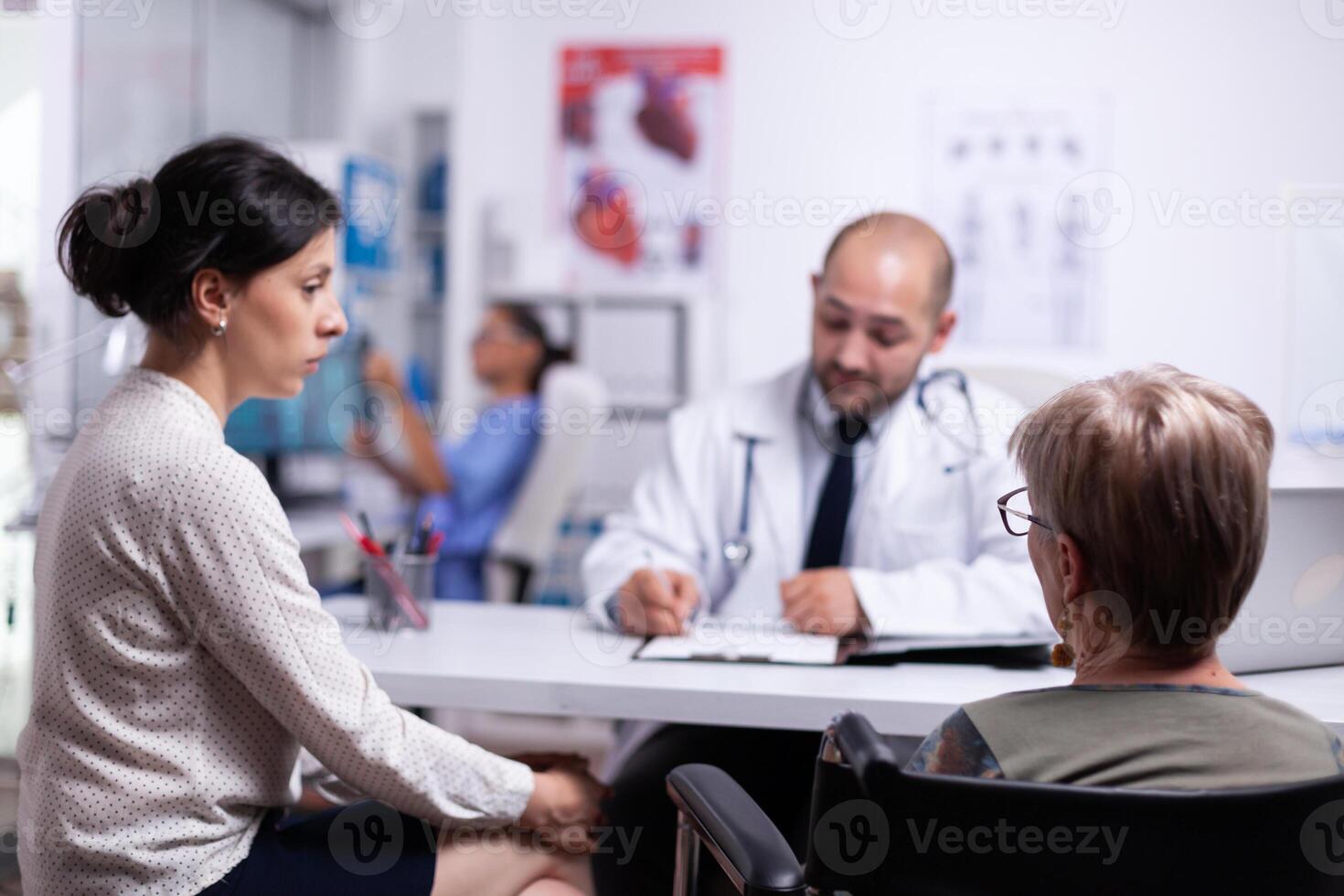 Doughter and disabled mother sitting in wheelchair talking with doctor during consultation. Practitioner asking questions to mature patient filling disease history form, prescribing treatment photo