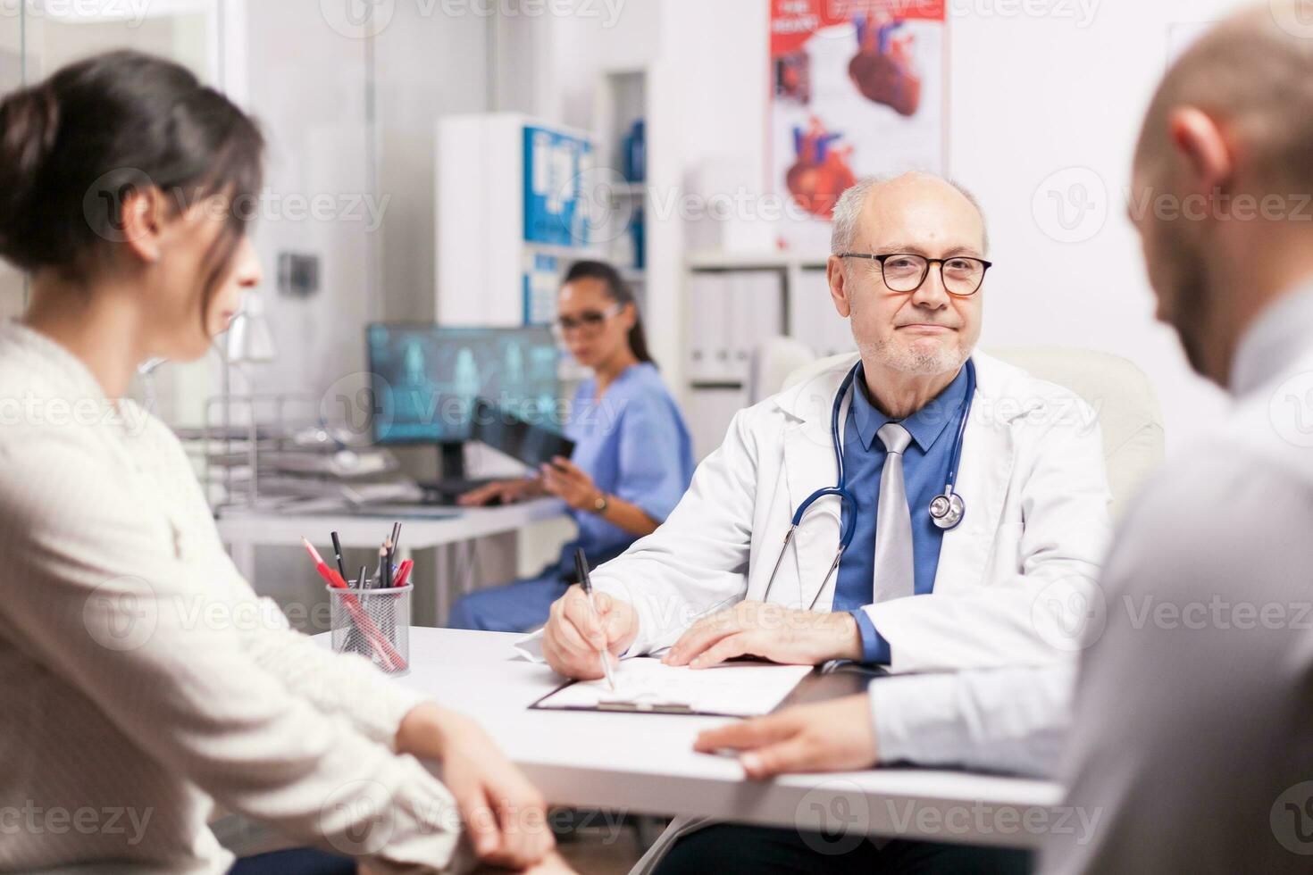 mayor médico facultativo tomando notas en portapapeles durante consulta de casado Pareja en hospital oficina. médico mirando a masculino paciente mientras vistiendo blanco Saco y estetoscopio. foto