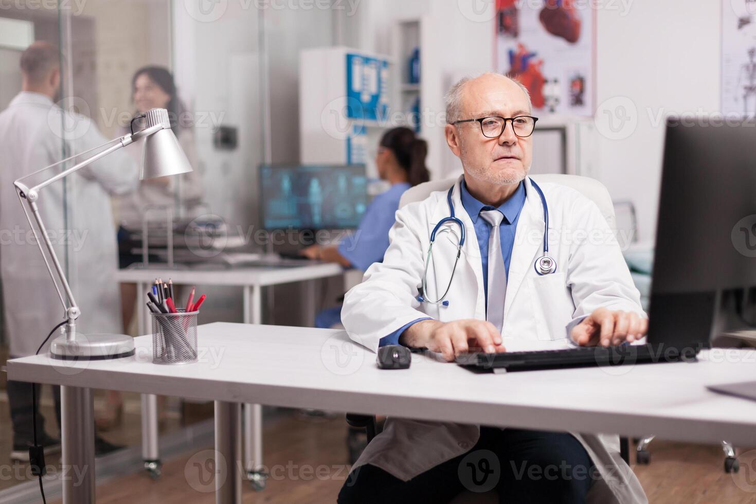 Senior man doctor with grey hair working on computer while young colleague is with a patient on hospital corridor and nurse checking analysis report. photo