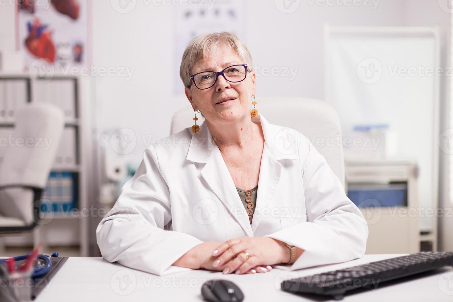 Medic with grey hair wearing coat in clinic cabinet sitting at desk. photo