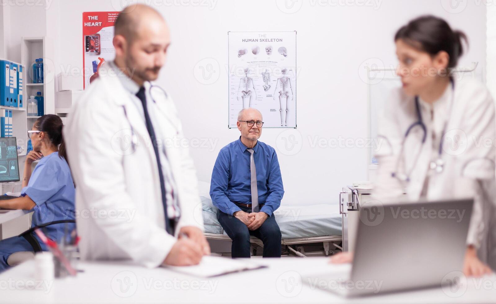 Two specialists doctors discussing senior patient diagnosis in hospital office. Scared mature man waiting medic expertise for his illness. Nurse working on laptop. photo