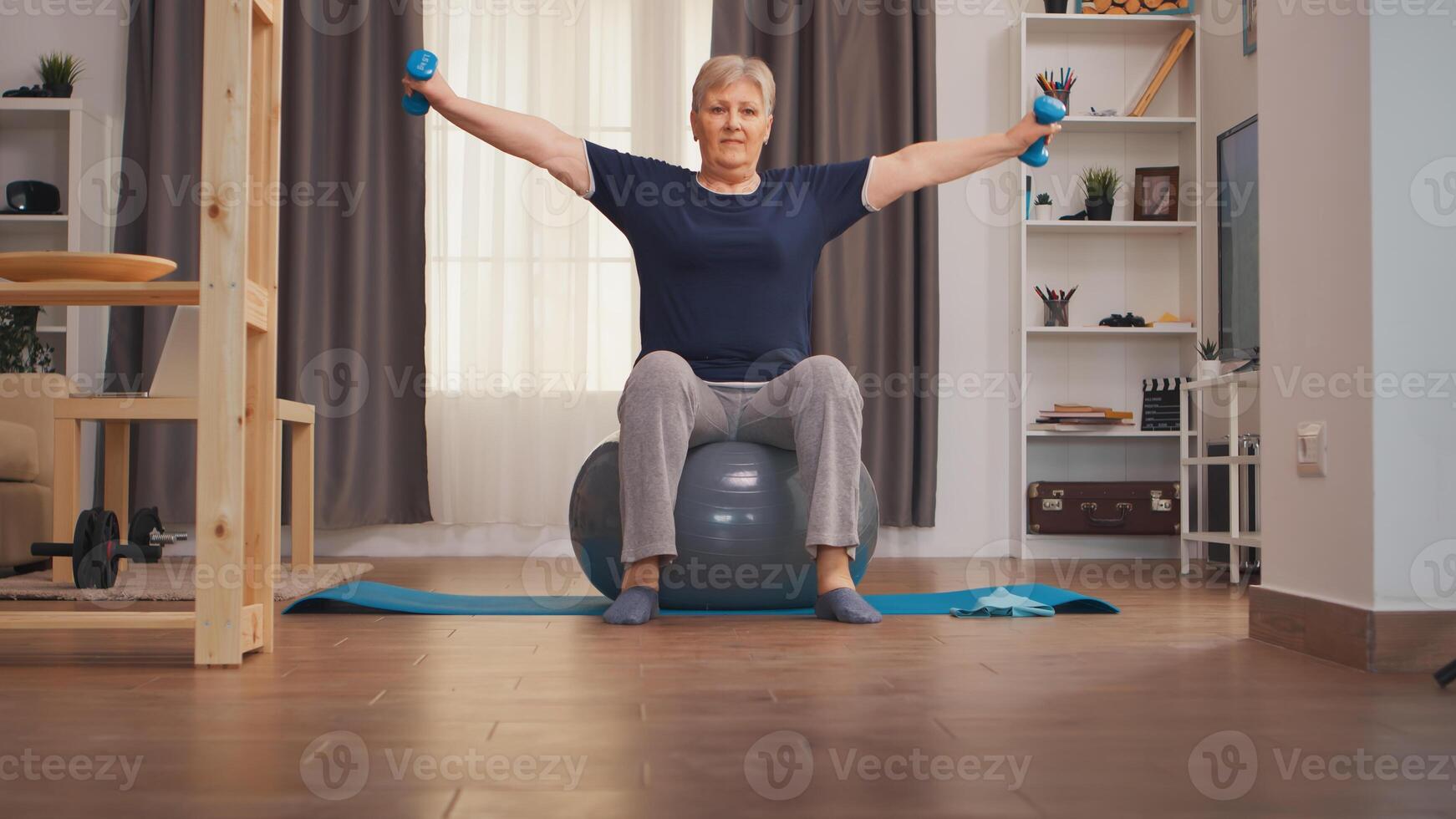 Senior woman training shoulders with dumbbells sitting on swiss ball. Old woman lifting training healthy lifestyle sport fitness workout at home with weights dumbbell activity photo