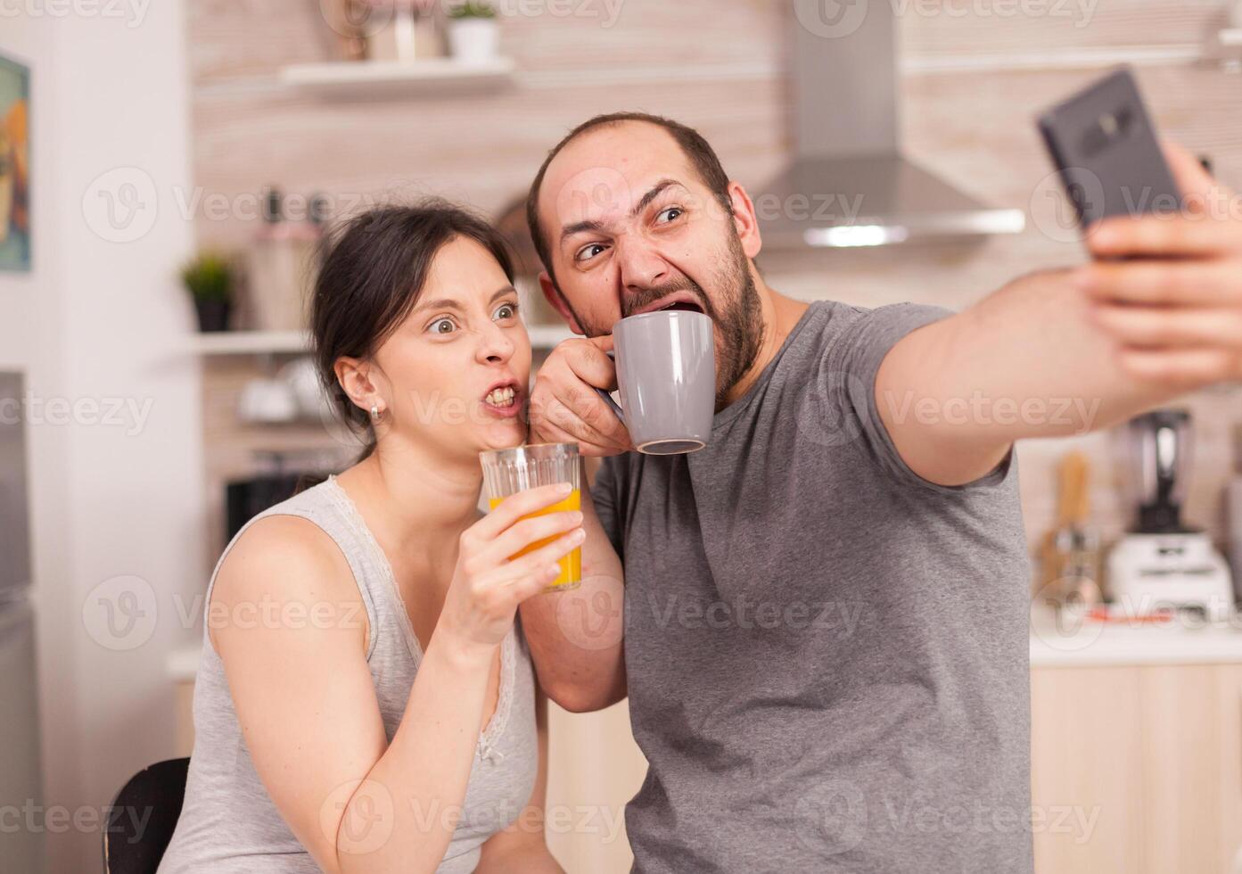 Happy lovers taking selfies with phone in the morning drinking orange juice and coffee. Joyful married husband and wife making funny faces while taking a photo during breakfast in kitchen.