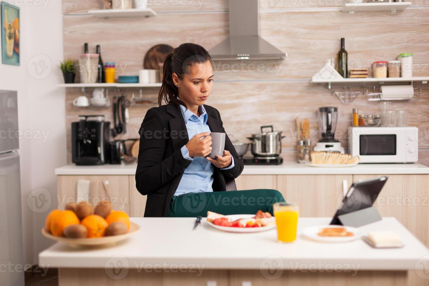 Young freelancer drinking coffee in the kitchen while using a tablet for reading the rapport. Business woman reading the last news online before going to work, using modern technology in the kitchen while eating a healthy meal. photo