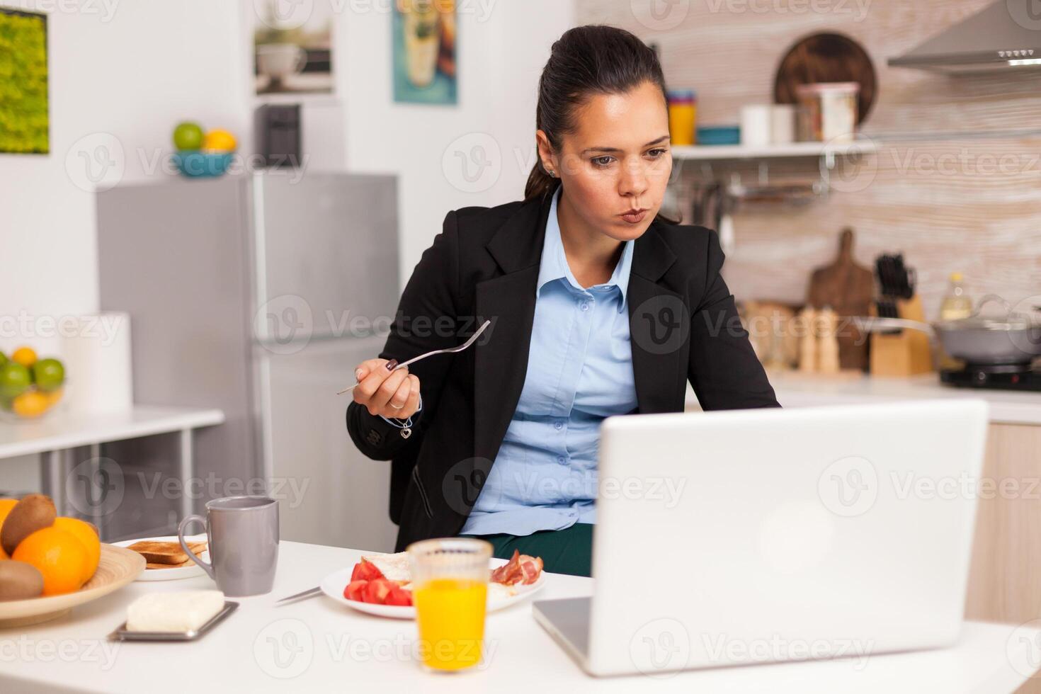 Businessperson eating breakfast while working from home. Concentrated business woman in the morning multitasking in the kitchen before going to the office, stressful way of life, career and goals to meet. photo